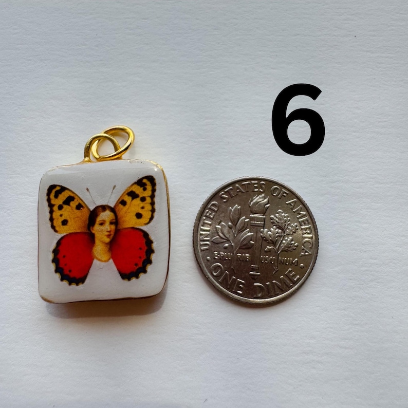 May include: A square pendant with a butterfly design. The pendant has a gold-colored bail and a white background. The butterfly has yellow, orange, and red wings. A dime is next to the pendant for size comparison.