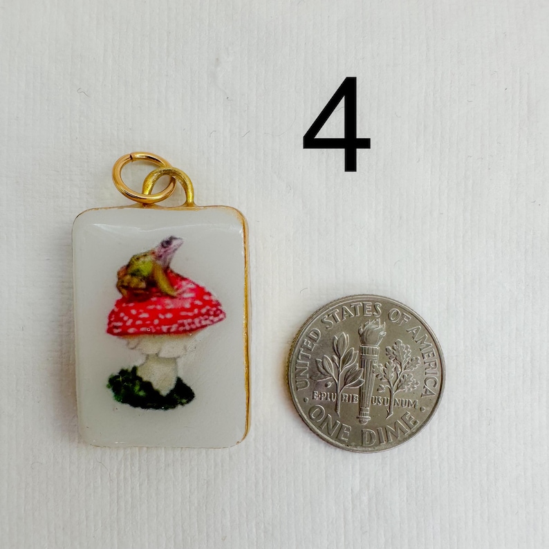 May include: A rectangular pendant with a gold-colored frame features a painted mushroom design with a red cap and white spots. A small frog sits atop the mushroom. A dime is next to the pendant for size comparison.