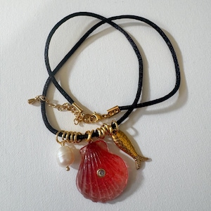 May include: A black cord necklace featuring a gold-tone metal clasp and extender chain. The pendant arrangement includes a white pearl bead, a translucent red seashell charm with a small embedded crystal, and a gold-tone metal fish charm.