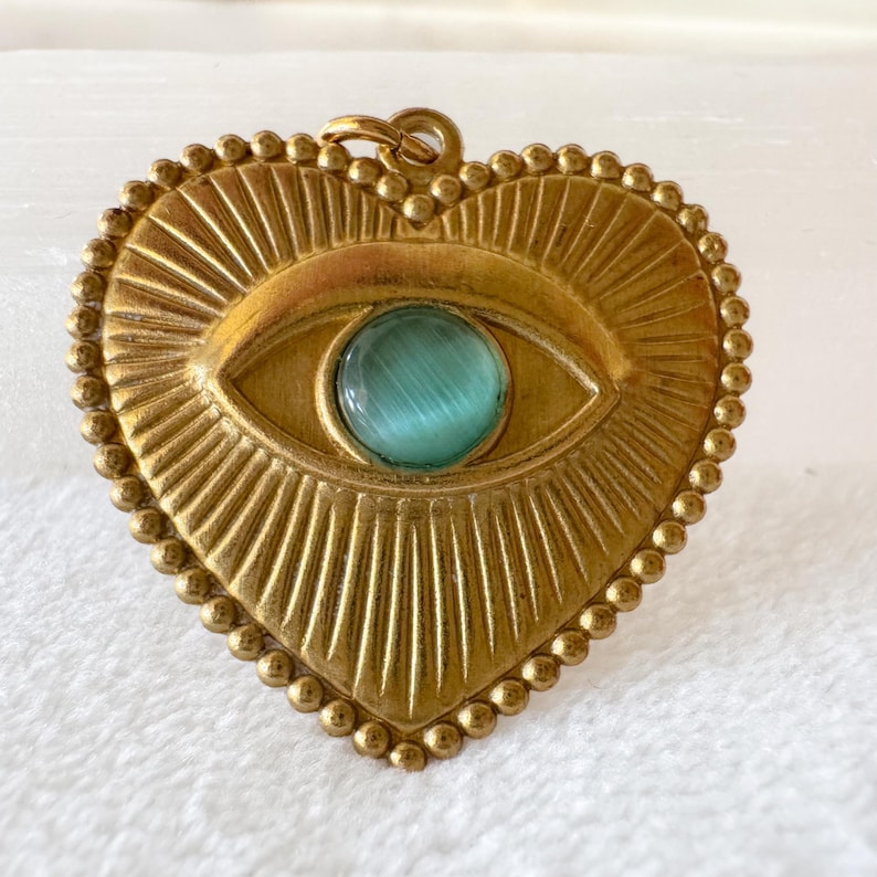 May include: A heart-shaped pendant with an eye design. The pendant is gold-colored with a teal-colored eye center. The eye is surrounded by sunburst rays and a beaded border. The pendant has a small loop at the top for hanging.