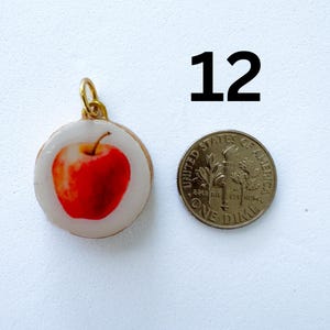 K&ouml;nnte beinhalten: Ein runder Anh&auml;nger mit einer roten Apfelillustration auf wei&szlig;em Hintergrund, eingefasst in einen goldfarbenen Metallrand mit &Ouml;se. Die Zahl 12 ist schwarz &uuml;ber dem Anh&auml;nger aufgedruckt, der neben einer US-amerikanischen 10-Cent-M&uuml;nze liegt.