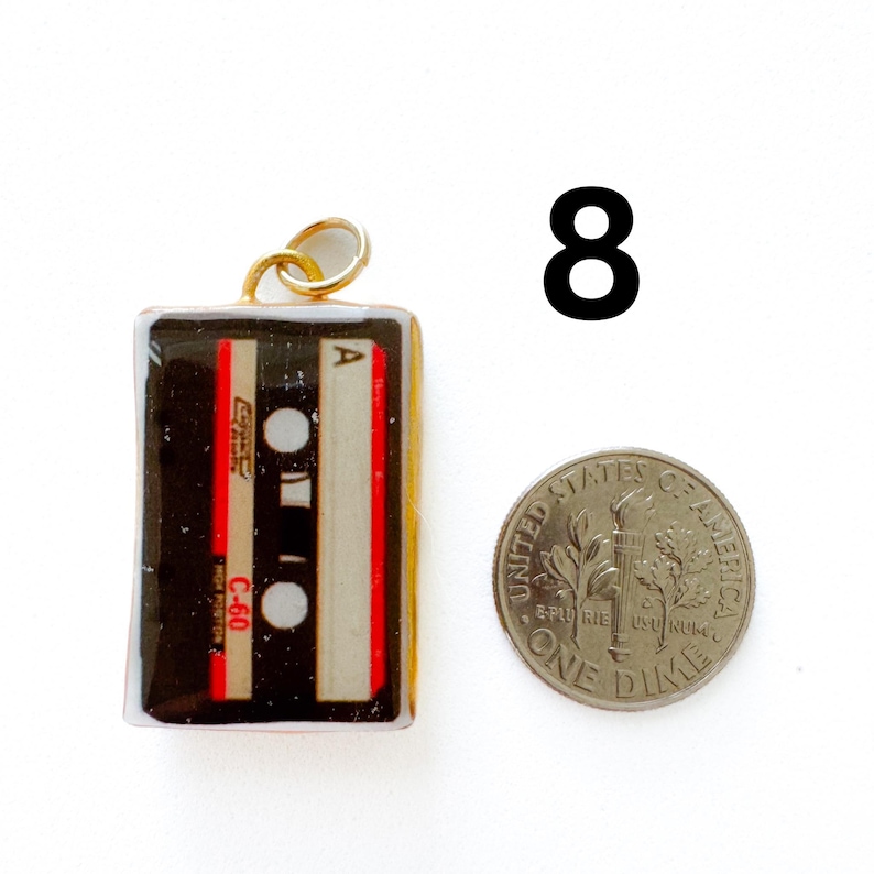 K&ouml;nnte beinhalten: Ein rechteckiger Anh&auml;nger in Form einer Vintage-Kassette mit schwarzen, wei&szlig;en und roten Details sowie der Aufschrift C-60 und A. Ein goldfarbener Biegering ist oben befestigt. Eine US-amerikanische Zehn-Cent-M&uuml;nze liegt daneben.