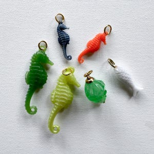 May include: A collection of six small charms featuring four seahorses in green, dark blue, orange, and light yellow, one white fish, and one faceted green bead, each attached to a small gold-toned metal jump ring for hanging or jewelry attachment.