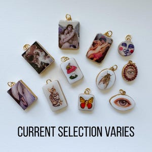 May include: A collection of rectangular and oval pendants with gold-colored bails. The pendants feature various images, including a cat, a butterfly, a mushroom, and classical art. The text "CURRENT SELECTION VARIES" is at the bottom.