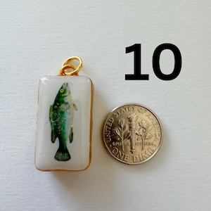 May include: A rectangular pendant featuring a green fish illustration on a white background with a gold-toned metal border and jump ring. The pendant is positioned next to a United States one dime coin for scale, with the number 10 displayed above.