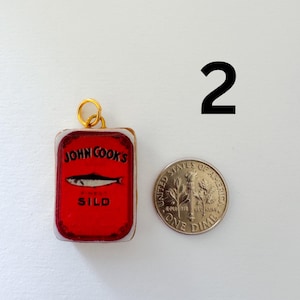 May include: A rectangular charm featuring a red John Cook's Finest Sild sardine tin graphic with a gold jump ring, positioned next to a United States one dime coin for scale. The charm displays a black fish illustration and black text on red.