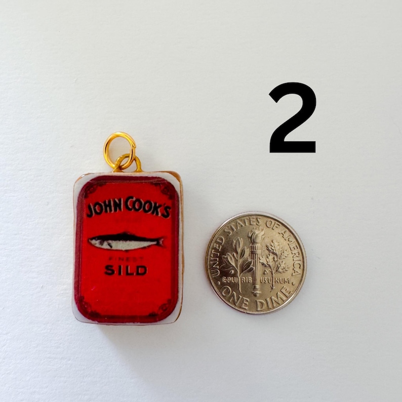 May include: A rectangular charm featuring a red John Cook's Finest Sild sardine tin graphic with a gold jump ring, positioned next to a United States one dime coin for scale. The charm displays a black fish illustration and black text on red.