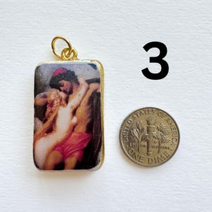 May include: A rectangular pendant with a gold-colored frame and a loop at the top. The pendant features a classical painting of two figures embracing. A dime is next to the pendant, and the number "3" is in the background.
