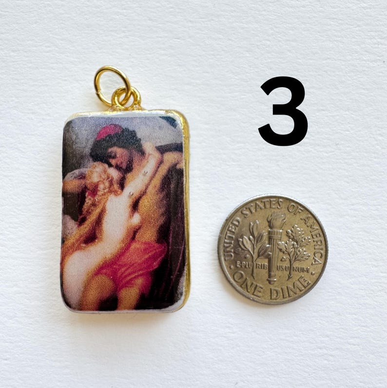 May include: A rectangular pendant with a gold-colored frame and a loop at the top. The pendant features a classical painting of two figures embracing. A dime is next to the pendant, and the number "3" is in the background.
