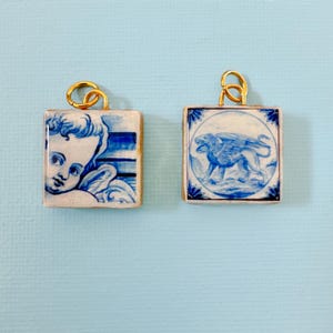 May include: Two square pendants with gold-colored loops. One pendant features a blue and white image of an angel, the other a winged lion. The pendants have a gold-colored border and are set against a light blue background.