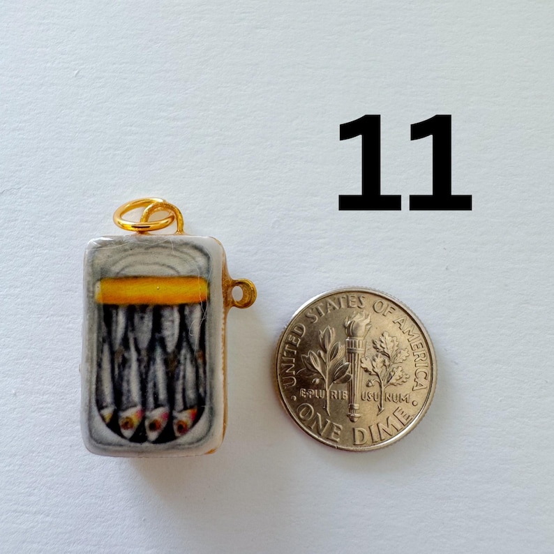 May include: A rectangular pendant shaped like an open tin of sardines with a yellow band, featuring a gold-tone jump ring and a side loop. The pendant is shown next to a United States dime for scale, with the number 11 printed in black above.