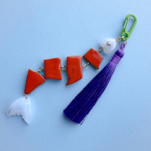 Peut inclure: Un porte-clés artisanal avec un motif de poisson stylisé. Le poisson est composé de segments rectangulaires orange avec des bords dorés, reliés par de petites chaînes dorées. Il a une queue blanche translucide et un pompon violet. Un mousqueton vert et une perle blanche en forme de cœur complètent le design.