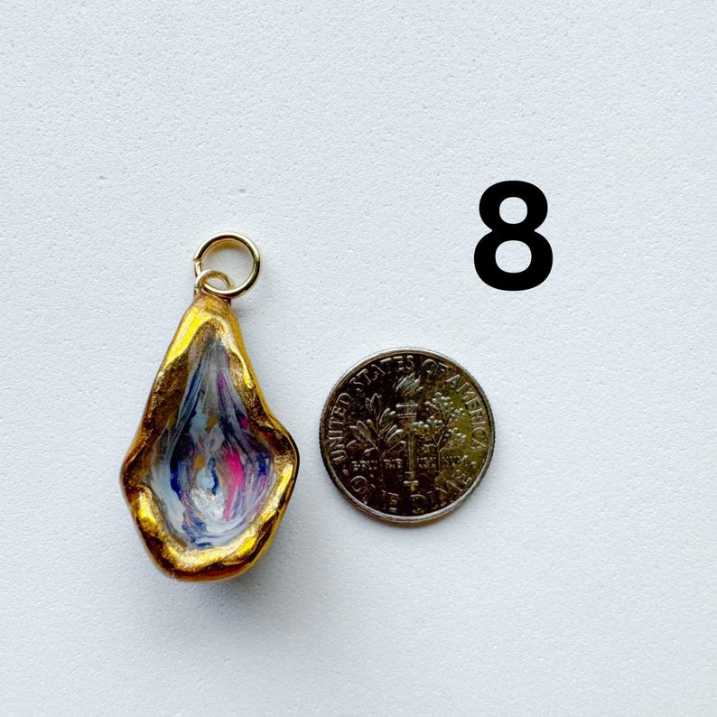 May include: A teardrop-shaped pendant with a gold-painted rim and a swirling interior of white, blue, and pink colors. A small metal jump ring is attached to the top. A United States dime is placed next to the pendant for scale, and the number 8.