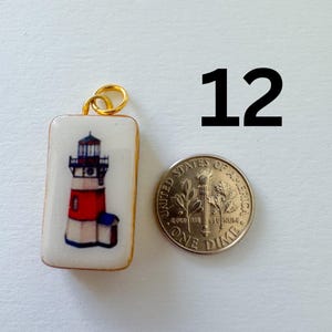 May include: A rectangular white pendant featuring a lighthouse illustration with red and blue accents, topped with a gold jump ring. Next to the pendant is a United States dime for scale, and the number 12 is printed in black in the upper right.