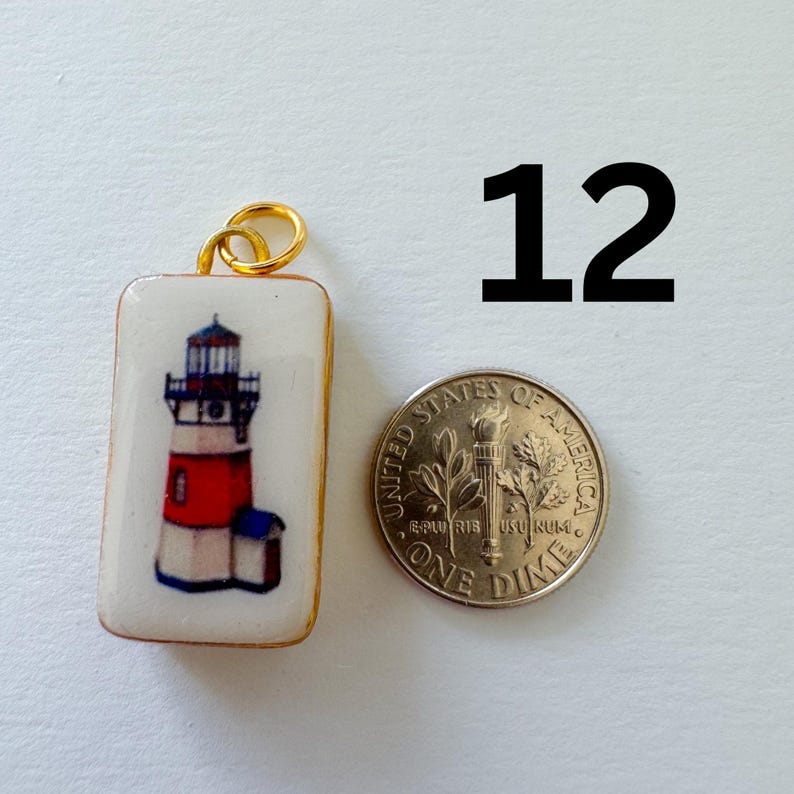 May include: A rectangular white pendant featuring a lighthouse illustration with red and blue accents, topped with a gold jump ring. Next to the pendant is a United States dime for scale, and the number 12 is printed in black in the upper right.