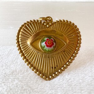 May include: A gold-colored heart-shaped pendant with an eye design. The eye features a colorful floral center. The pendant has a textured surface with a beaded border and a loop for hanging. The pendant is approximately 2 inches wide.