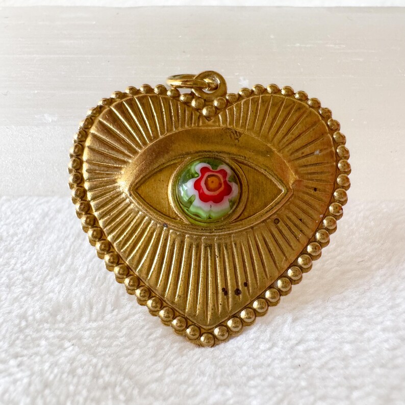 May include: A gold-colored heart-shaped pendant with an eye design. The eye features a colorful floral center. The pendant has a textured surface with a beaded border and a loop for hanging. The pendant is approximately 2 inches wide.