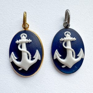 May include: Two oval pendants featuring a white anchor relief on a dark blue background. One pendant is set in a gold-toned metal frame with a matching gold-toned bail, while the other is set in a silver-toned metal frame with a silver-toned bail.