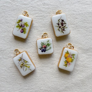 Può includere: Cinque ciondoli rettangolari bianchi con bordi color oro e un anello dorato nella parte superiore. Ogni ciondolo presenta un diverso design di frutta o botanico, tra cui fichi, olive e limoni. I ciondoli sono disposti su un tessuto neutro.