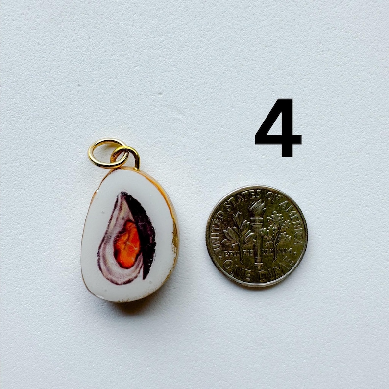 May include: A white, teardrop-shaped pendant featuring a detailed illustration of an open oyster with a bright orange center. The pendant has a gold-toned metal bail and is shown next to a United States dime for scale. The number 4 is printed nearby.