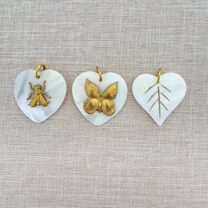 May include: Three white heart-shaped pendants with gold accents. The first pendant features a gold bee, the second a gold butterfly, and the third a carved leaf design.