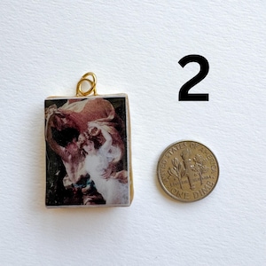 May include: A rectangular pendant with a gold-colored loop at the top, featuring a vintage artwork print in shades of red, white, and black. A dime is next to the pendant. The number "2" is in black.