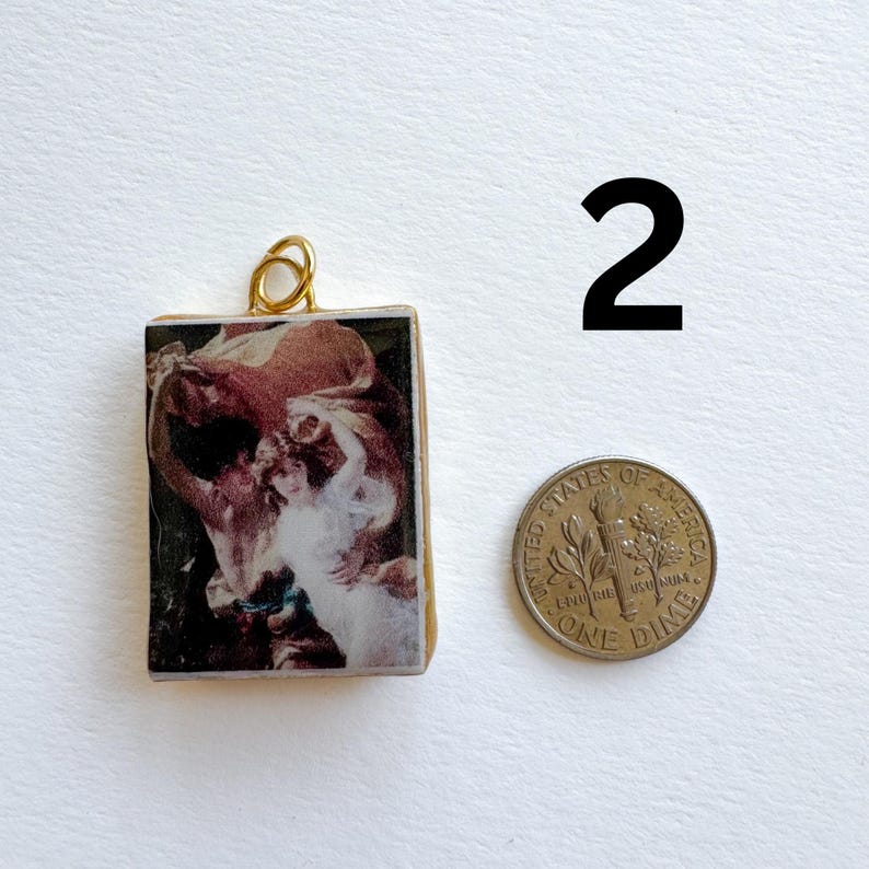 May include: A rectangular pendant with a gold-colored loop at the top, featuring a vintage artwork print in shades of red, white, and black. A dime is next to the pendant. The number "2" is in black.