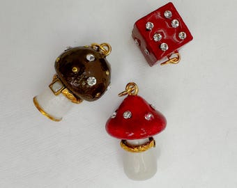 Könnte beinhalten: Drei dekorative Anhänger: ein brauner Pilz, ein roter Pilz und ein roter Würfel. Jeder Anhänger ist mit Strasssteinen und goldfarbenen Akzenten verziert. Die Pilzanhänger haben weiße Stiele, und der Würfel hat weiße Punkte.