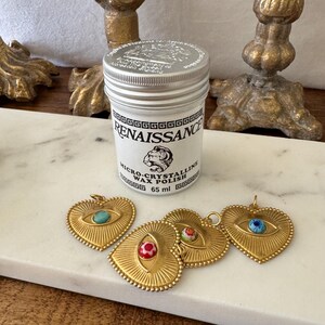 May include: A white jar of Renaissance Micro-Crystalline Wax Polish, 65 ml, with a silver lid, sits near four gold-colored heart-shaped pendants with eye designs. The pendants have turquoise, red, and blue accents.
