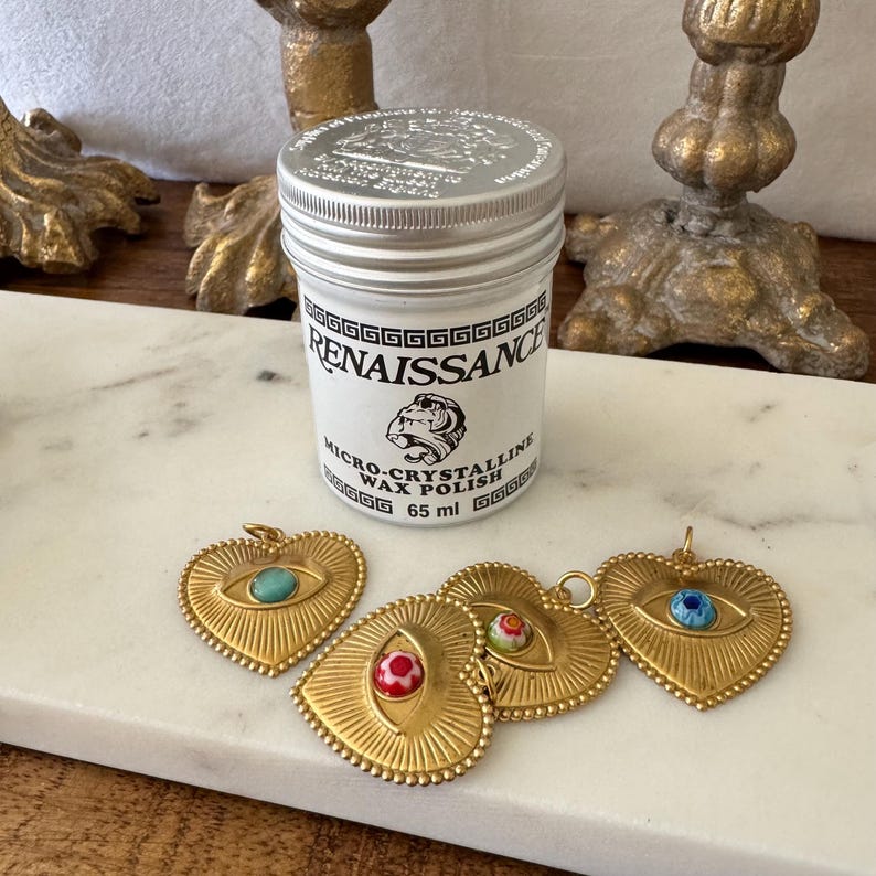 May include: A white jar of Renaissance Micro-Crystalline Wax Polish, 65 ml, with a silver lid, sits near four gold-colored heart-shaped pendants with eye designs. The pendants have turquoise, red, and blue accents.