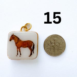 K&ouml;nnte beinhalten: Ein quadratischer Anh&auml;nger mit einer braunen Pferdeillustration auf wei&szlig;em Hintergrund mit einem goldfarbenen Metallrahmen und einem Biegering, platziert neben einer amerikanischen Zehn-Cent-M&uuml;nze zum Gr&ouml;&szlig;envergleich, mit der Zahl 15 in Schwarz dar&uuml;ber.