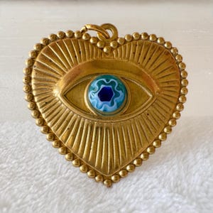 May include: A heart-shaped pendant with a gold-colored finish. The pendant features an eye design with a blue and white center. The border is adorned with small, round details. The eye is the focal point, with radiating lines extending from it.