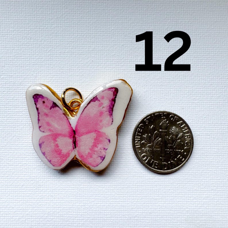 May include: A butterfly-shaped charm with a white base and pink watercolor-style wings, featuring a gold-toned metal edge and a small jump ring at the top. The charm is positioned next to a U.S. dime for scale, with the number 12 printed above.