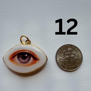May include: A pendant with a detailed eye design, featuring shades of red and purple, set in a white oval frame with a gold-colored border. A dime is next to the pendant for size comparison. The number "12" is above the dime.