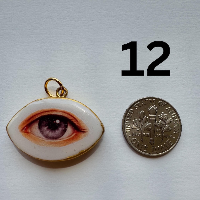 May include: A pendant with a detailed eye design, featuring shades of red and purple, set in a white oval frame with a gold-colored border. A dime is next to the pendant for size comparison. The number "12" is above the dime.