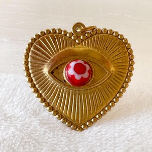 May include: A heart-shaped pendant with a gold-colored frame and a central eye design. The eye features a red and white glass bead. The pendant has a beaded border and a loop for hanging.
