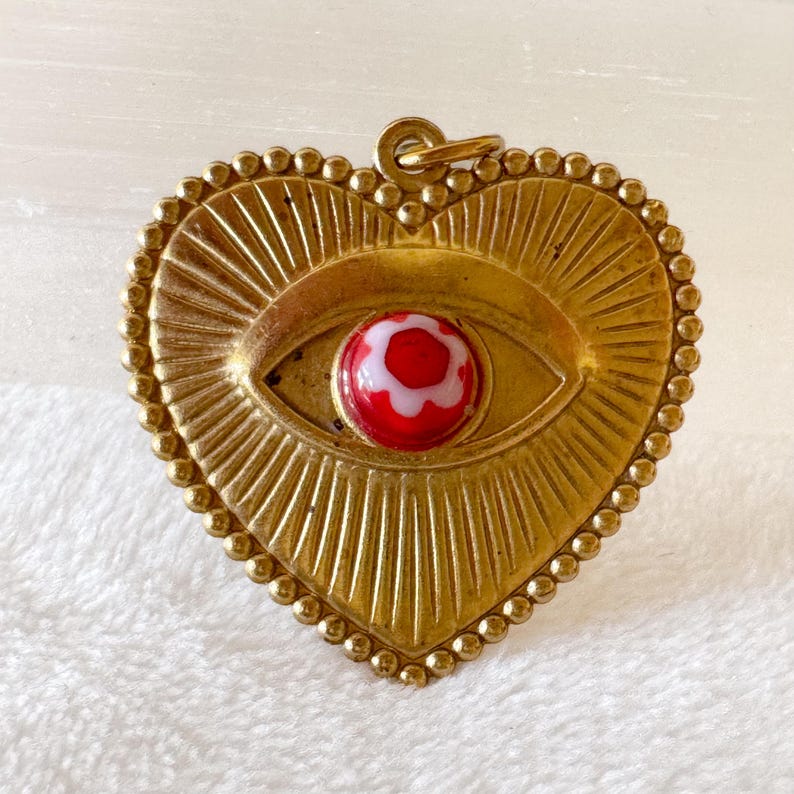 May include: A heart-shaped pendant with a gold-colored frame and a central eye design. The eye features a red and white glass bead. The pendant has a beaded border and a loop for hanging.