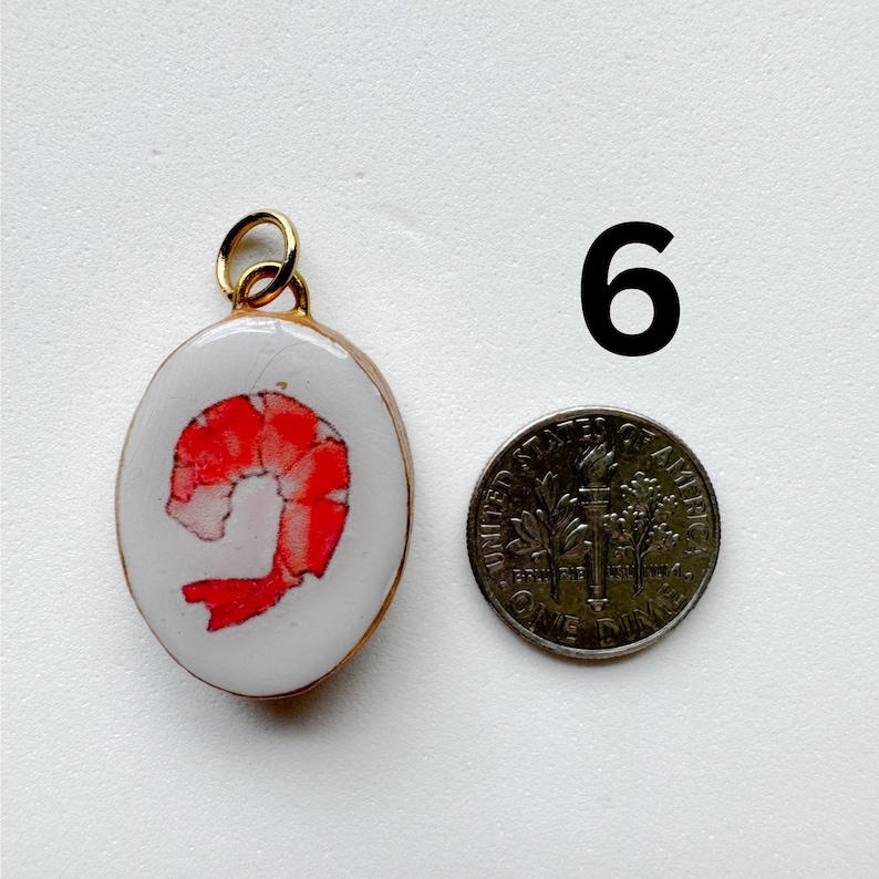 May include: An oval-shaped pendant featuring a red shrimp illustration on a white background with a gold-toned metal rim and jump ring. The pendant is positioned next to a United States dime for scale, with the number 6 printed in black nearby.