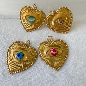 May include: Four gold-colored heart-shaped pendants with an eye design. Each pendant has a different colored eye: green, floral, blue, and red. The pendants have a textured sunburst design and a beaded border.