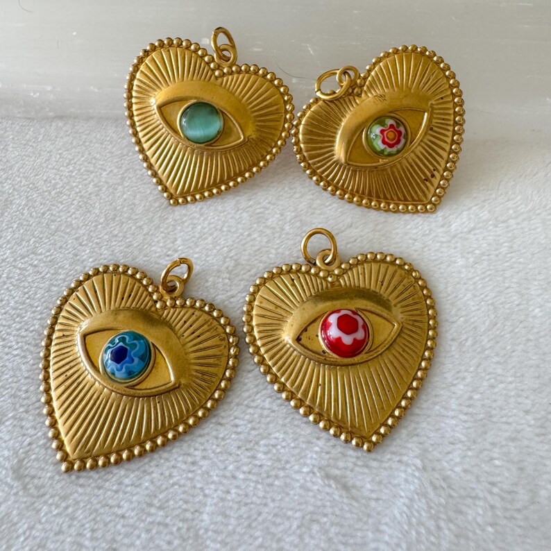 May include: Four gold-colored heart-shaped pendants with an eye design. Each pendant has a different colored eye: green, floral, blue, and red. The pendants have a textured sunburst design and a beaded border.