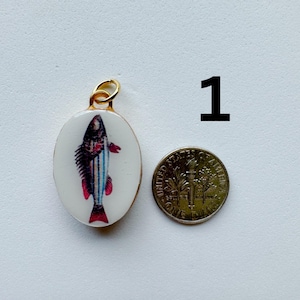 May include: An oval-shaped pendant with a white background featuring a colorful fish illustration with blue stripes and red fins. The pendant has a gold-toned metal bail and is shown next to a United States dime for scale, labeled with the number 1.