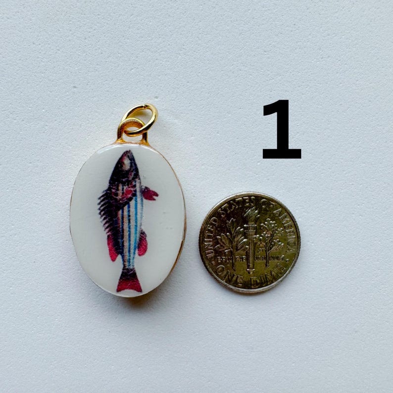 May include: An oval-shaped pendant with a white background featuring a colorful fish illustration with blue stripes and red fins. The pendant has a gold-toned metal bail and is shown next to a United States dime for scale, labeled with the number 1.