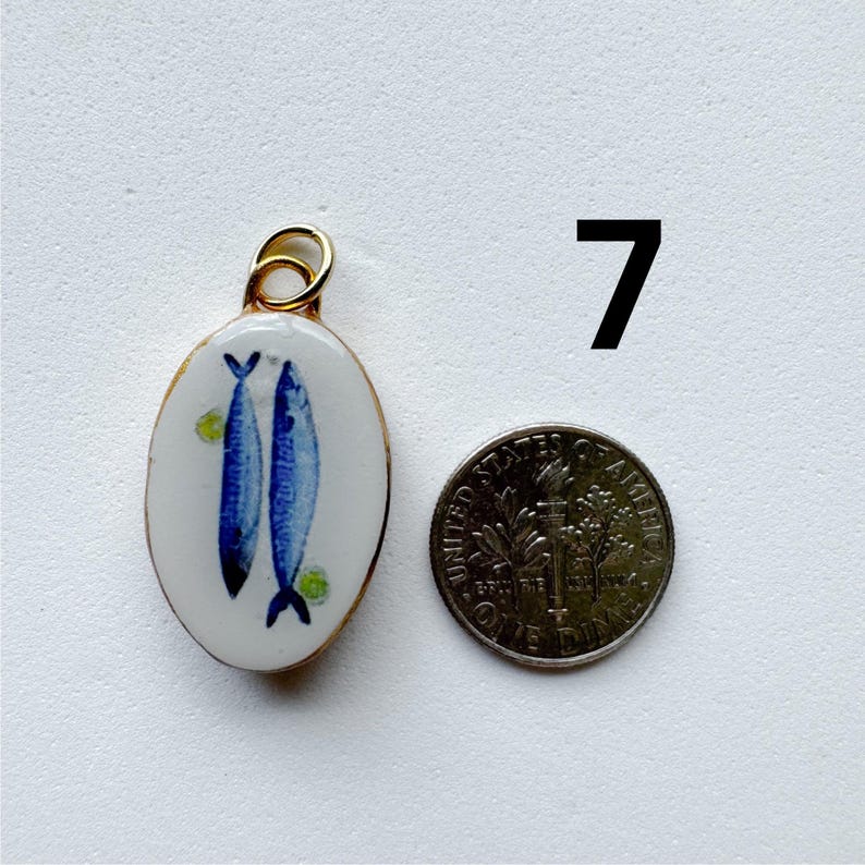 May include: An oval-shaped pendant with a white enamel surface featuring two blue fish illustrations and small green accents, framed in gold metal with a top loop. Next to it is a United States dime for scale and the number 7.