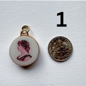 May include: A round white ceramic pendant featuring a pink cameo-style profile portrait of a person with a gold-toned metal bail. The pendant is positioned next to a United States dime for scale, with the number 1 printed in black above them.