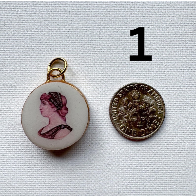 May include: A round white ceramic pendant featuring a pink cameo-style profile portrait of a person with a gold-toned metal bail. The pendant is positioned next to a United States dime for scale, with the number 1 printed in black above them.