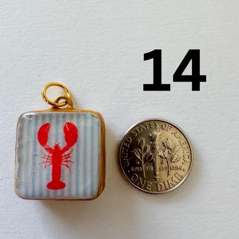 May include: A square pendant featuring a red lobster graphic on a light blue and white striped background, framed in gold with a jump ring at the top. The pendant is shown next to a United States one dime coin for scale, with the number 14 above.