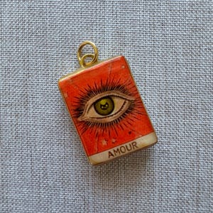 May include: A rectangular gold-toned pendant featuring a vibrant orange background with a detailed illustration of a single eye with long lashes and a green iris. The word AMOUR is printed in black capital letters at the bottom of the pendant.