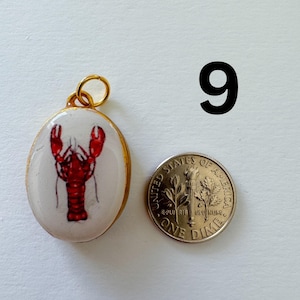 May include: An oval pendant with a gold-colored metal rim and a white center featuring a red lobster illustration. A gold jump ring is attached to the top. A United States one dime coin is placed next to the pendant for scale. The number 9 is visible.