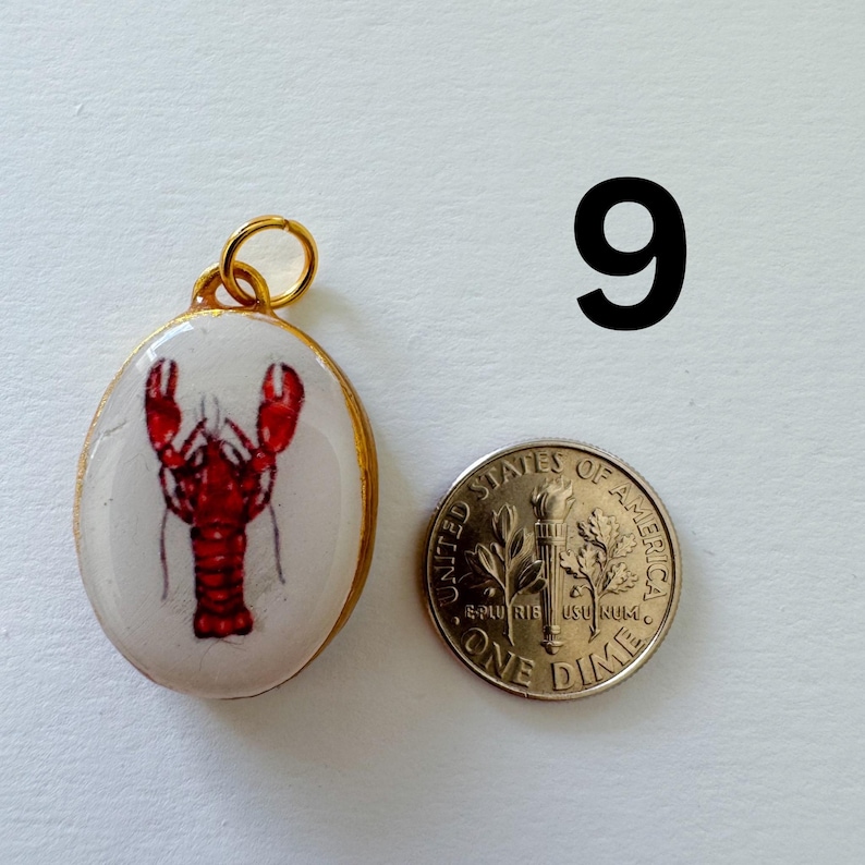 May include: An oval pendant with a gold-colored metal rim and a white center featuring a red lobster illustration. A gold jump ring is attached to the top. A United States one dime coin is placed next to the pendant for scale. The number 9 is visible.