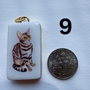 May include: A rectangular pendant with a gold-colored bail features a seated cat illustration with brown and black stripes on a white background. A dime is next to the pendant for size comparison.